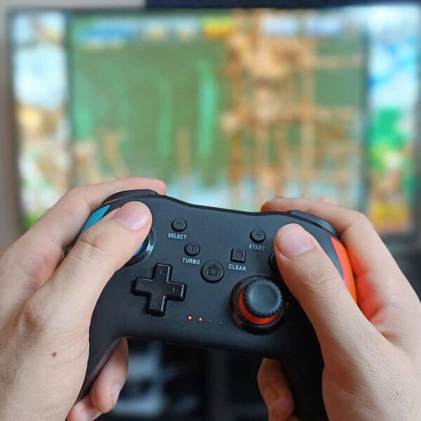 un mando inalambrico pro de color negro azul y rojo sujetado por la mano de un hombre y de fondo un videojuego retro